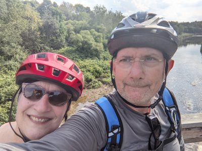 A couple in bike helmets posing with a landscape behind them including a stream and forest. The man is making a funny expression as he takes the selfie.